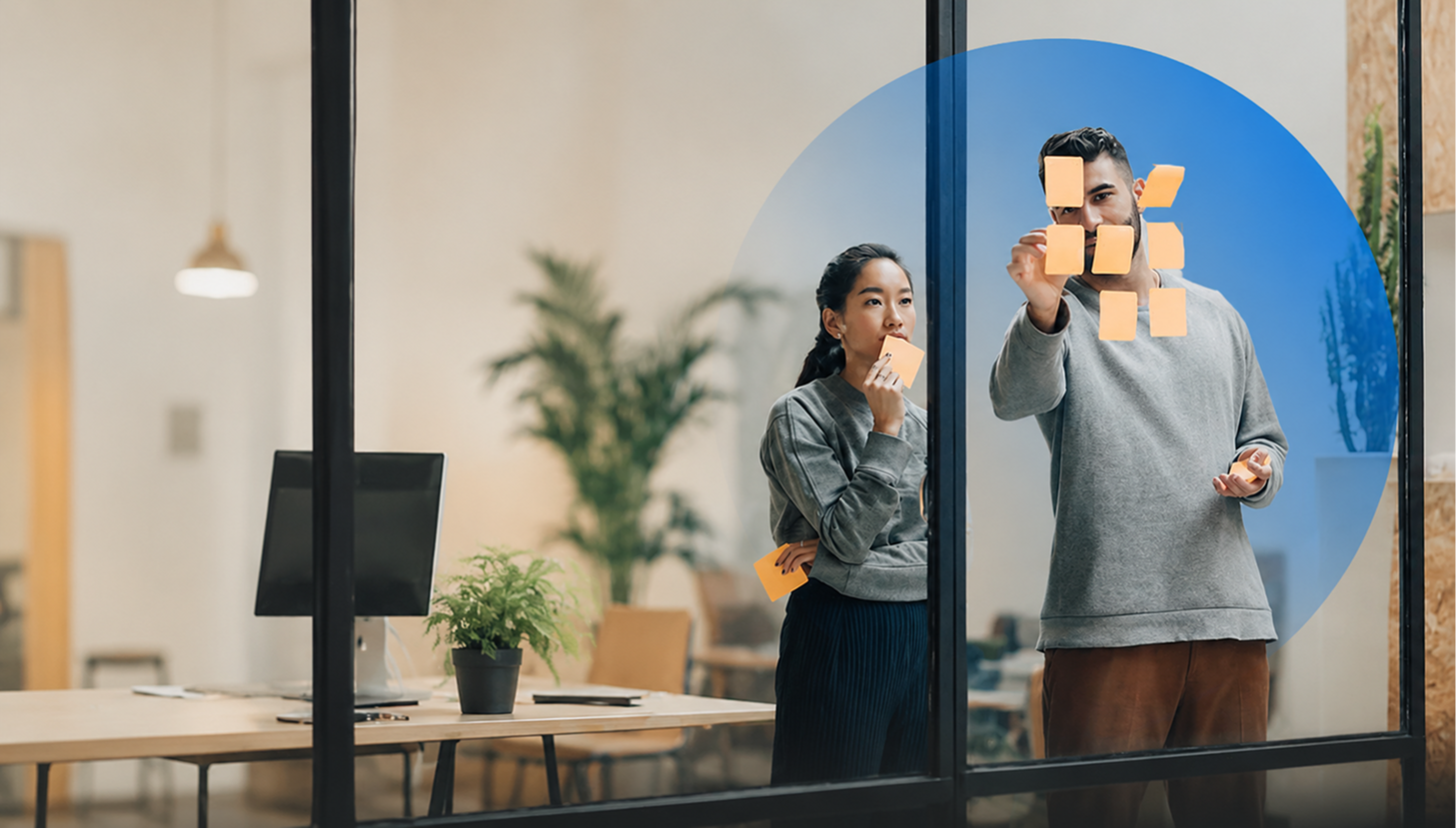 Two colleagues discuss sticky notes on a glass wall in a modern office.