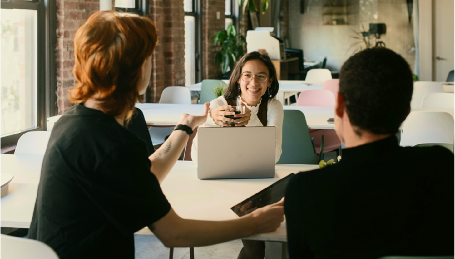 Three coworkers seated at a table with a laptop in a bright office.