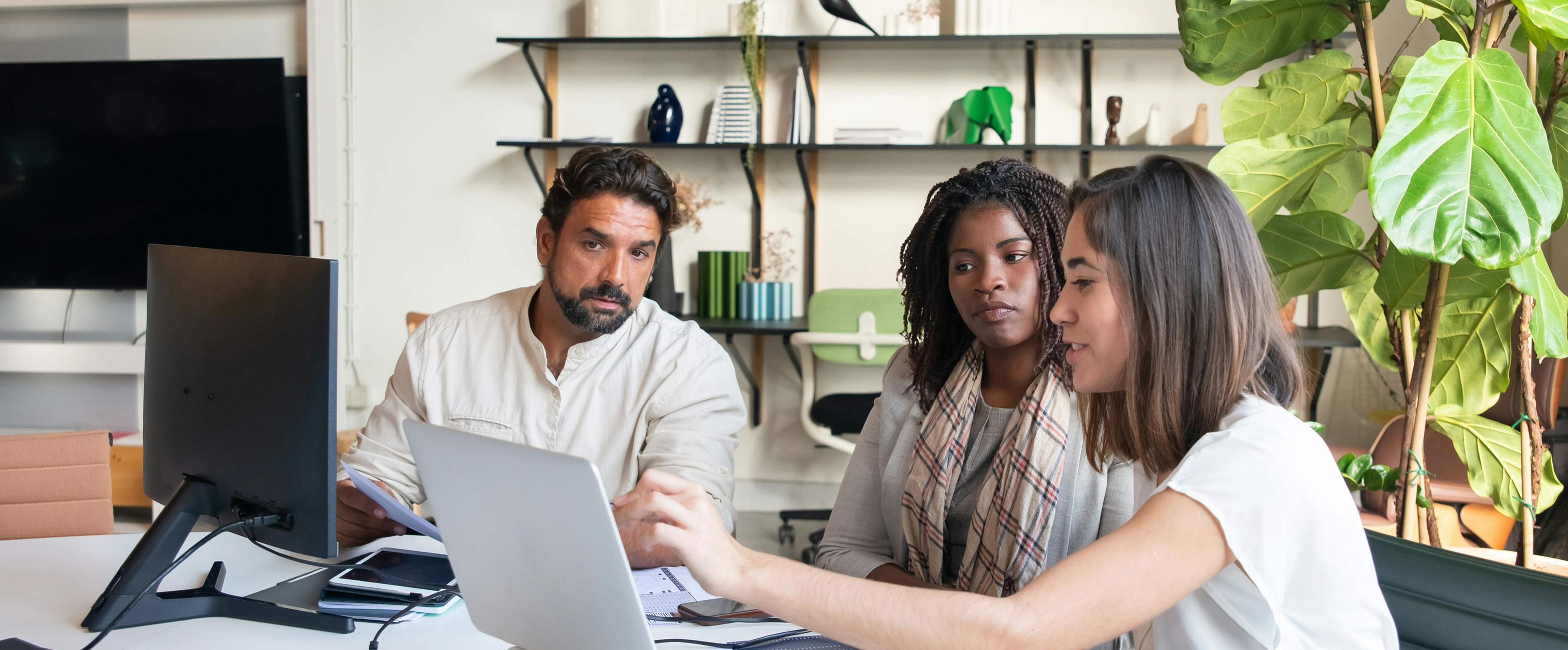 Three colleagues collaborating around laptops in a bright office with plants.
