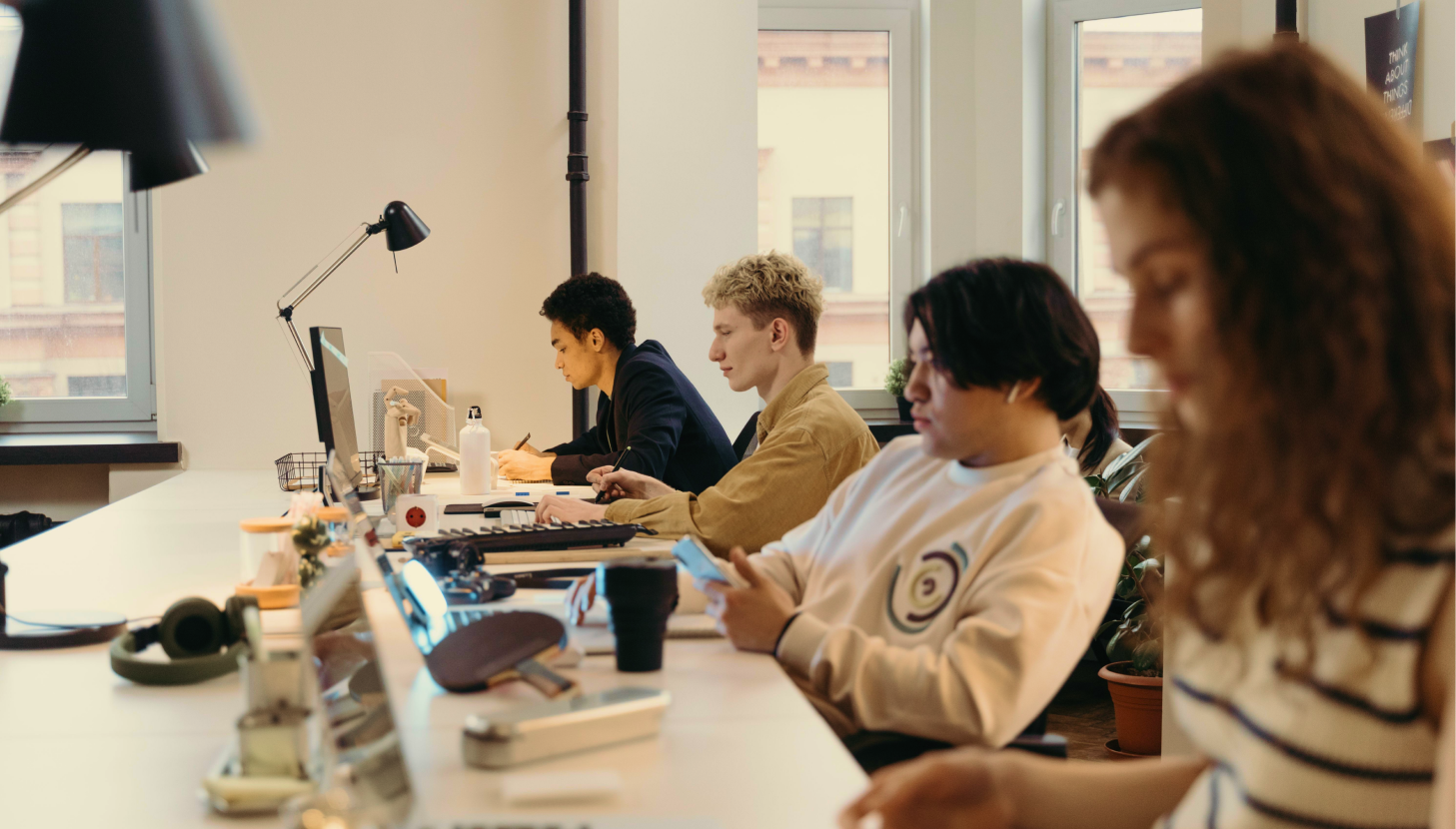 People sit at a shared desk working on laptops in a bright office space.