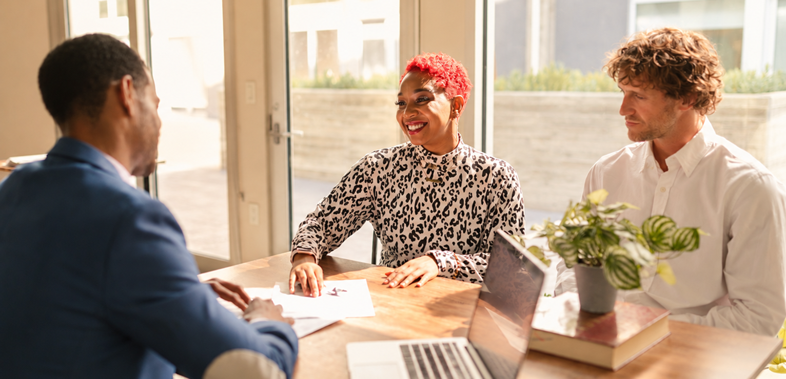 Three colleagues in a meeting at a table with a laptop and plant.