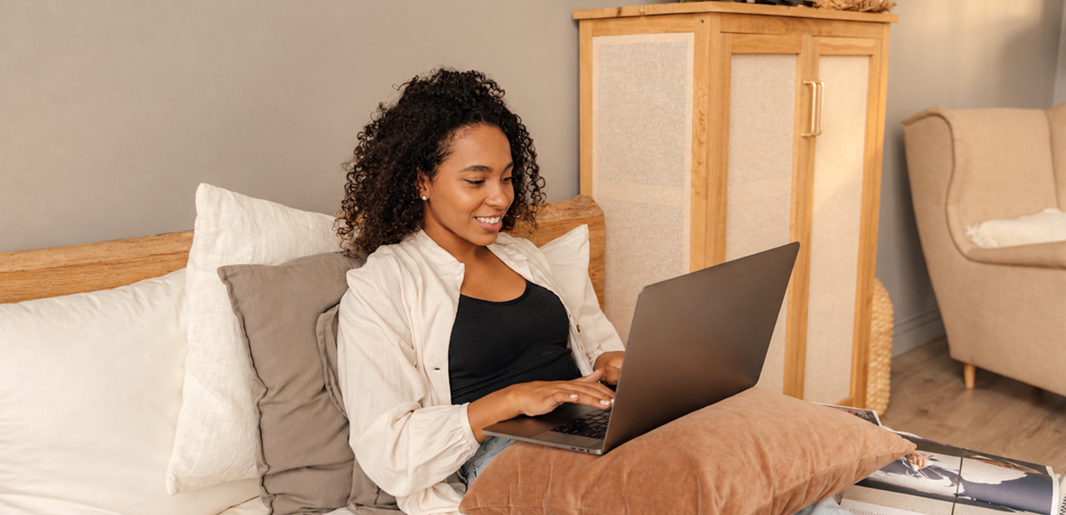 Woman sitting on bed smiling while using a laptop in a cozy room.