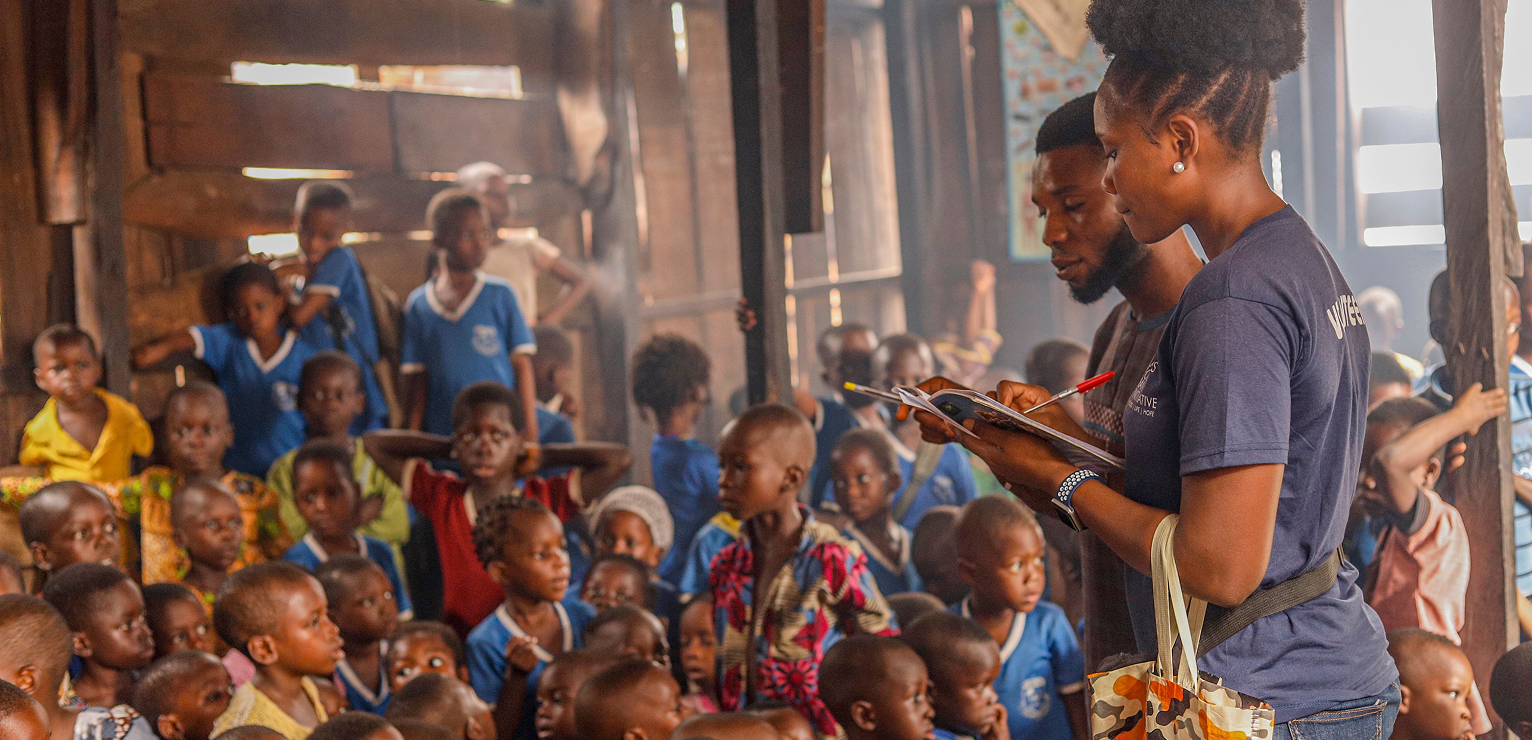 Man speaking to a large group of seated children outdoors, photograph.