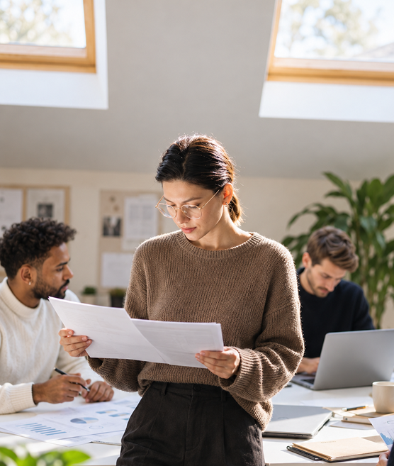 Woman reviewing documents in a bright office with colleagues at a desk.