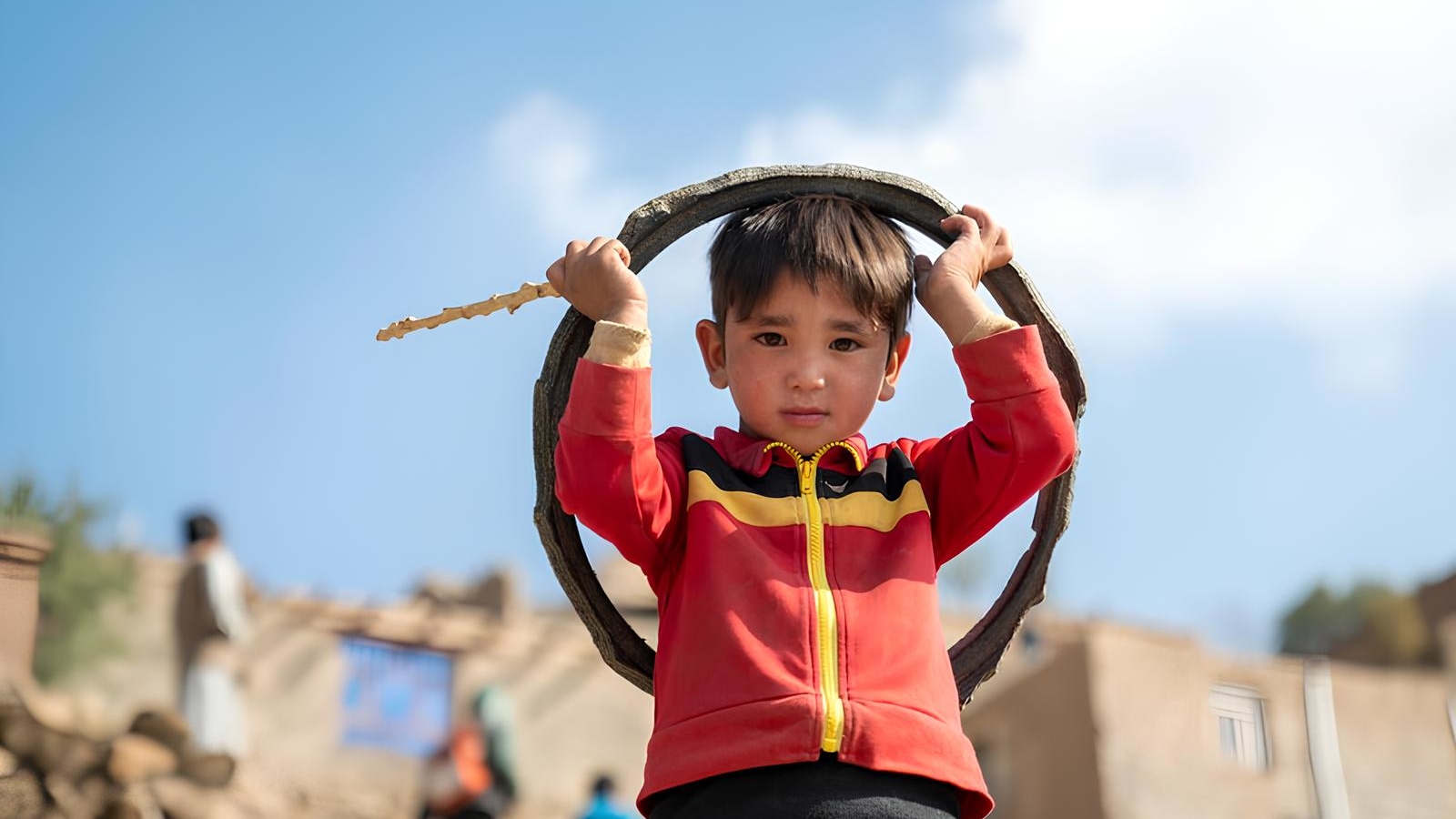 Young boy in vibrant red jacket plays with a hoop, holding a stick. Clear blue sky and rustic buildings in background, creating a joyful, playful mood.